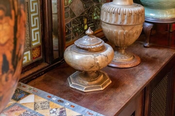 A small, ornate, lidded urn sits on a wooden shelf next to a larger vase and decorative stained glass.