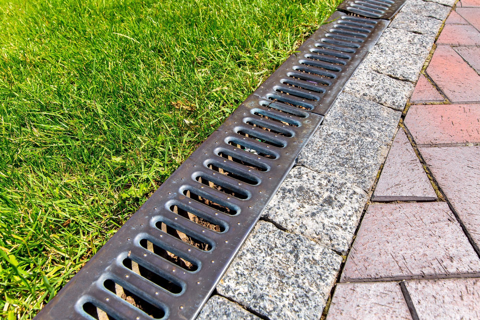 Metal drainage grate along a paved walkway and grassy lawn.