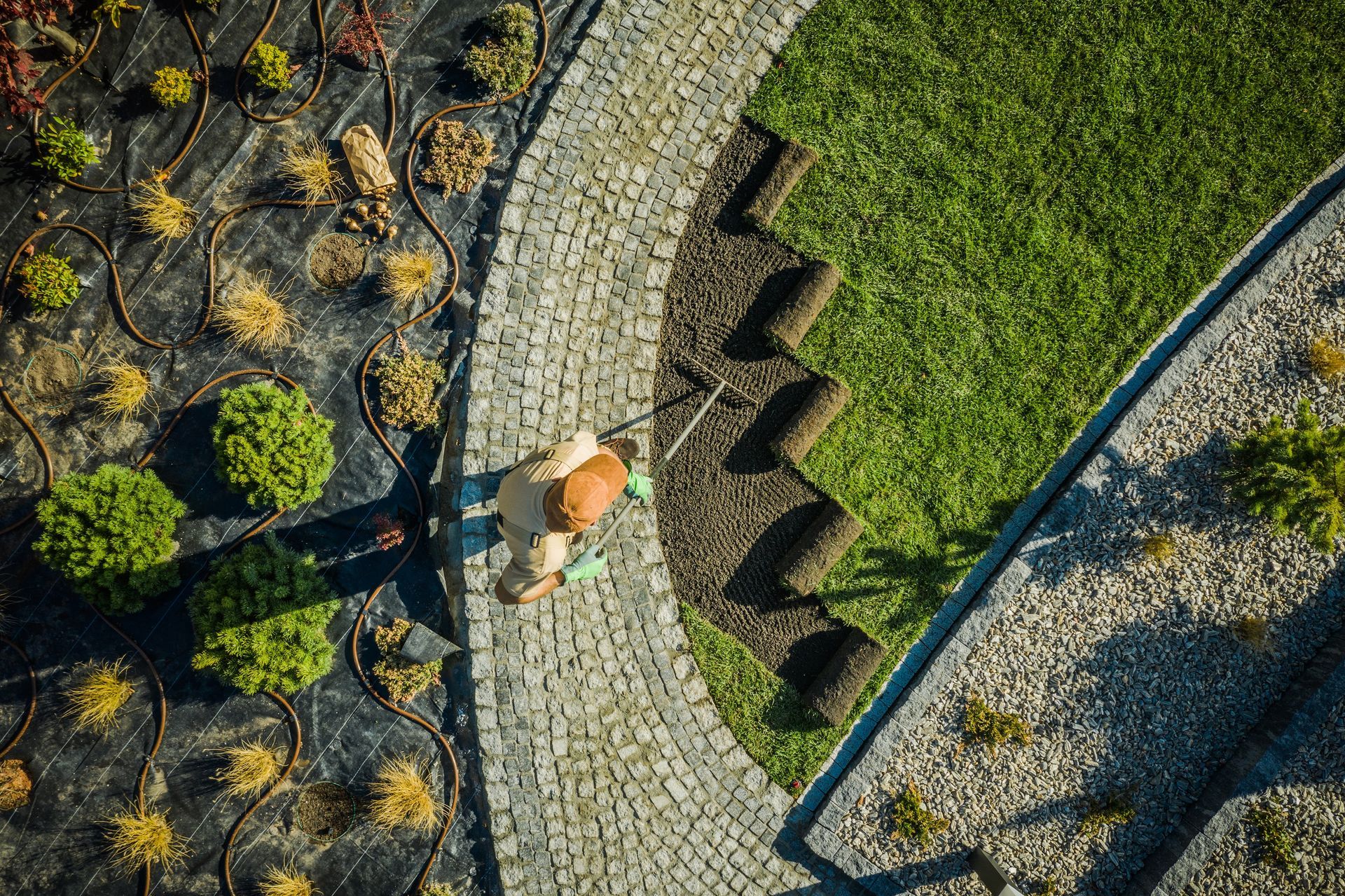 An overhead view of a person laying rolls of sod next to a cobblestone path in a garden with landscaped beds.