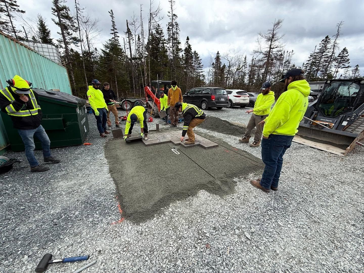 Workers in neon vests laying gravel surface outdoors near vehicles and a container.