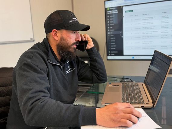 Man in black cap and jacket on a phone call, working on a laptop with a large monitor in the background.