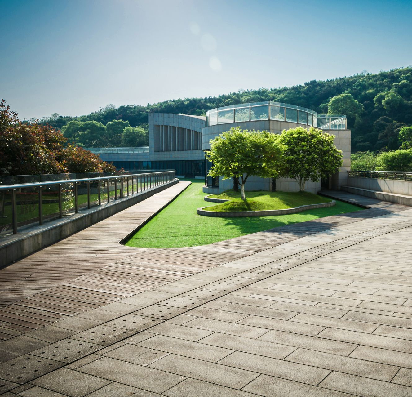 Brick walkway leading to modern building with glass roof, green lawn, trees, and hillside.