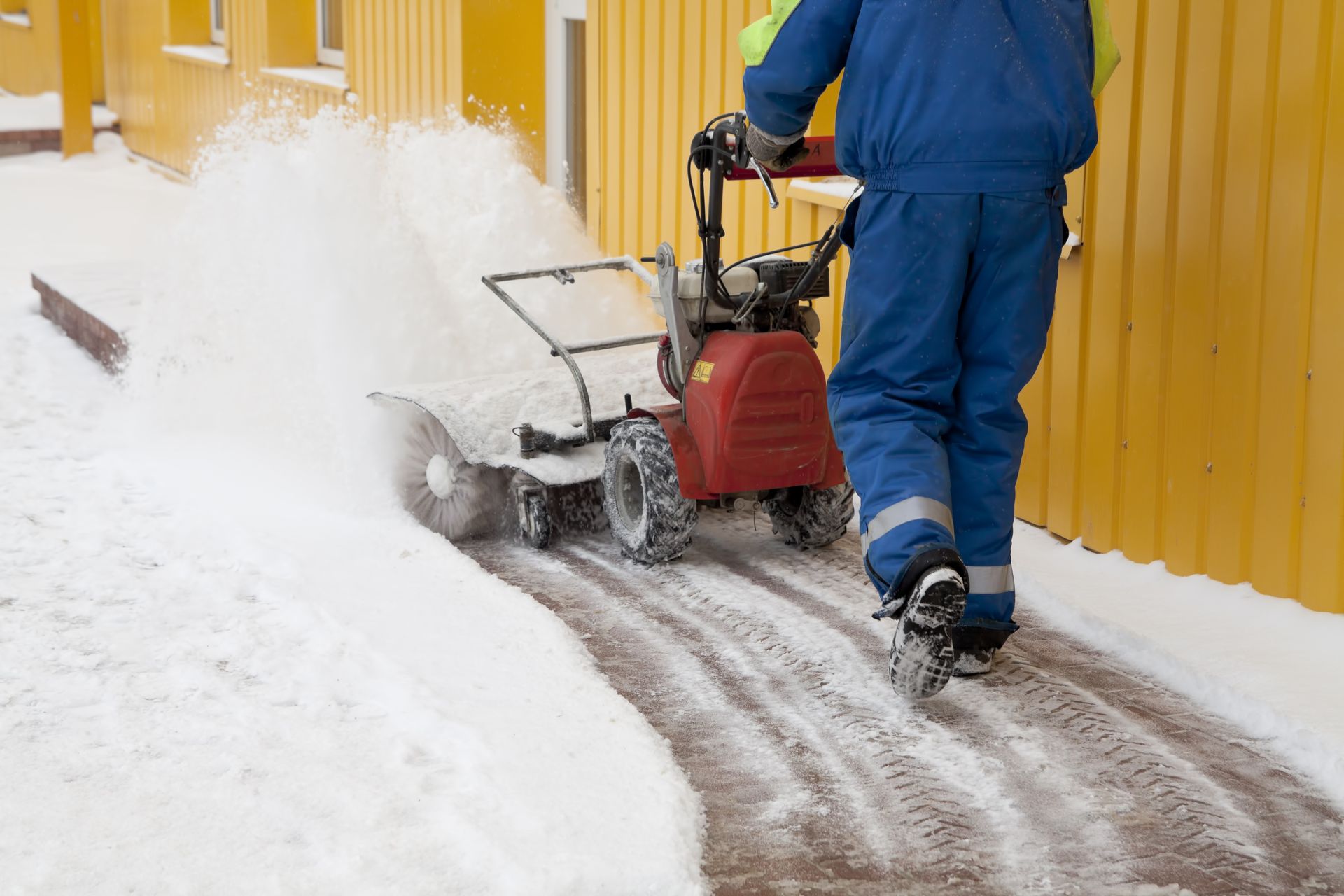 Person in blue overalls using a snowblower on a snowy sidewalk, next to a yellow building.