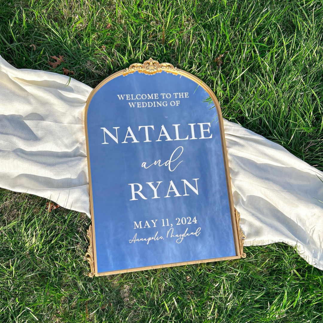 A blue wedding welcome sign for natalie and ryan is sitting on top of a lush green field.