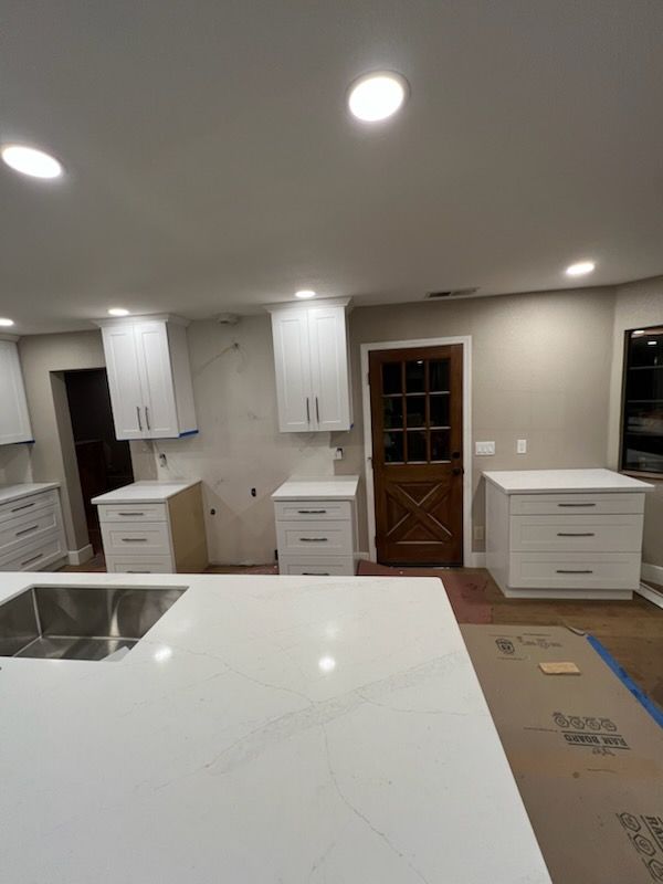 Kitchen remodel in progress: white cabinets installed, white countertop island, unfinished walls, brown door, and recessed lighting.