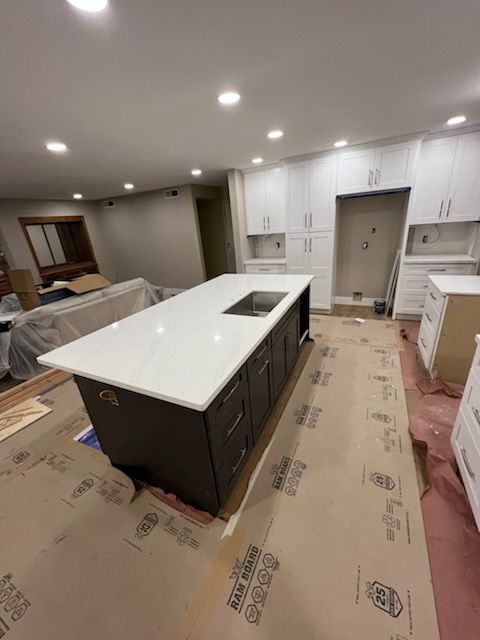 A kitchen renovation in progress with a dark brown island, white countertops, and unfinished cabinets against white walls. The floor is covered in protective cardboard.