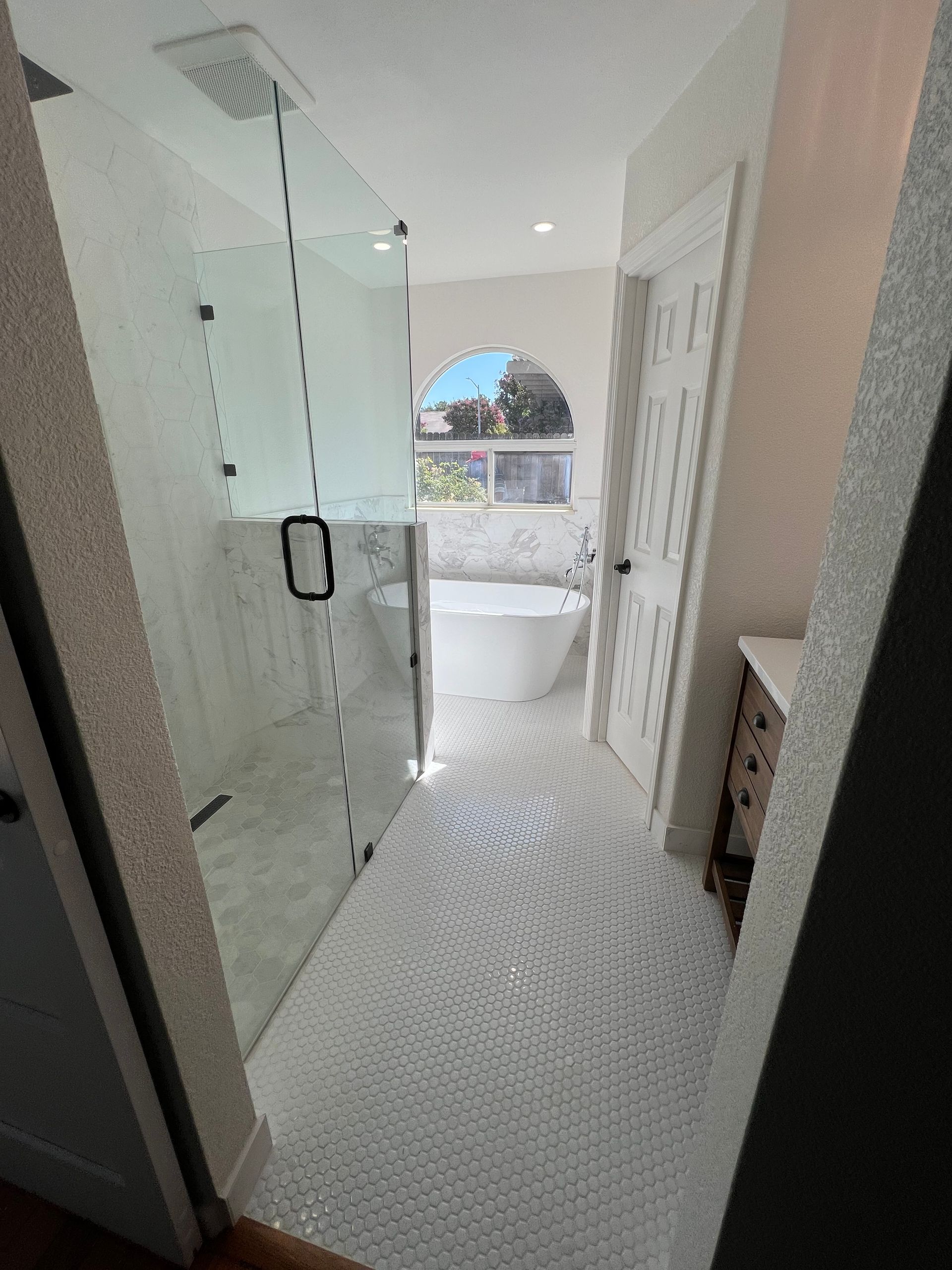Bright white bathroom with a glass shower, soaking tub, and white patterned floor. A doorway and cabinet are visible on the right.
