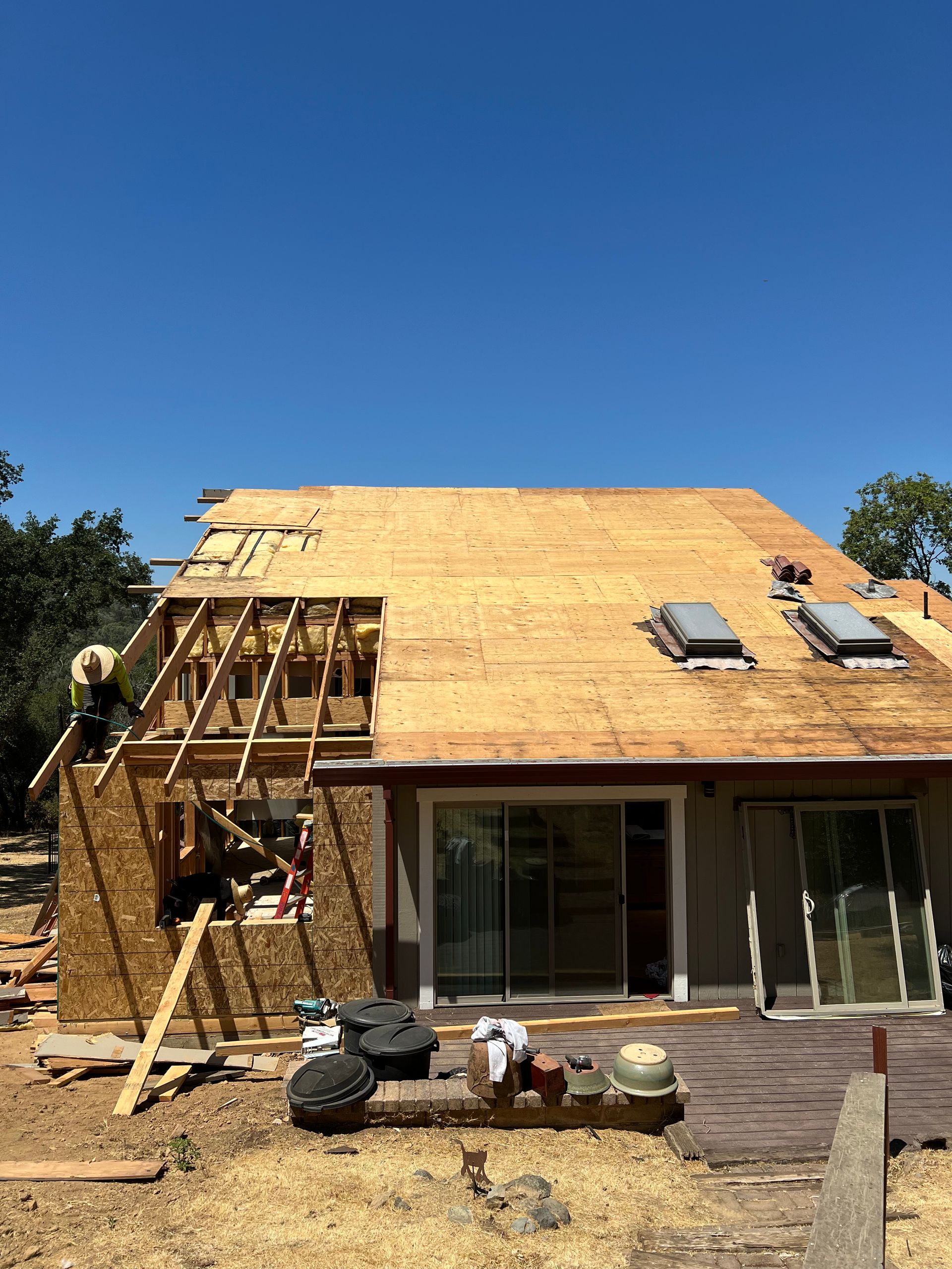 Construction workers on a roof removing old shingles. A partially built wooden structure with exposed beams, and bright blue sky.