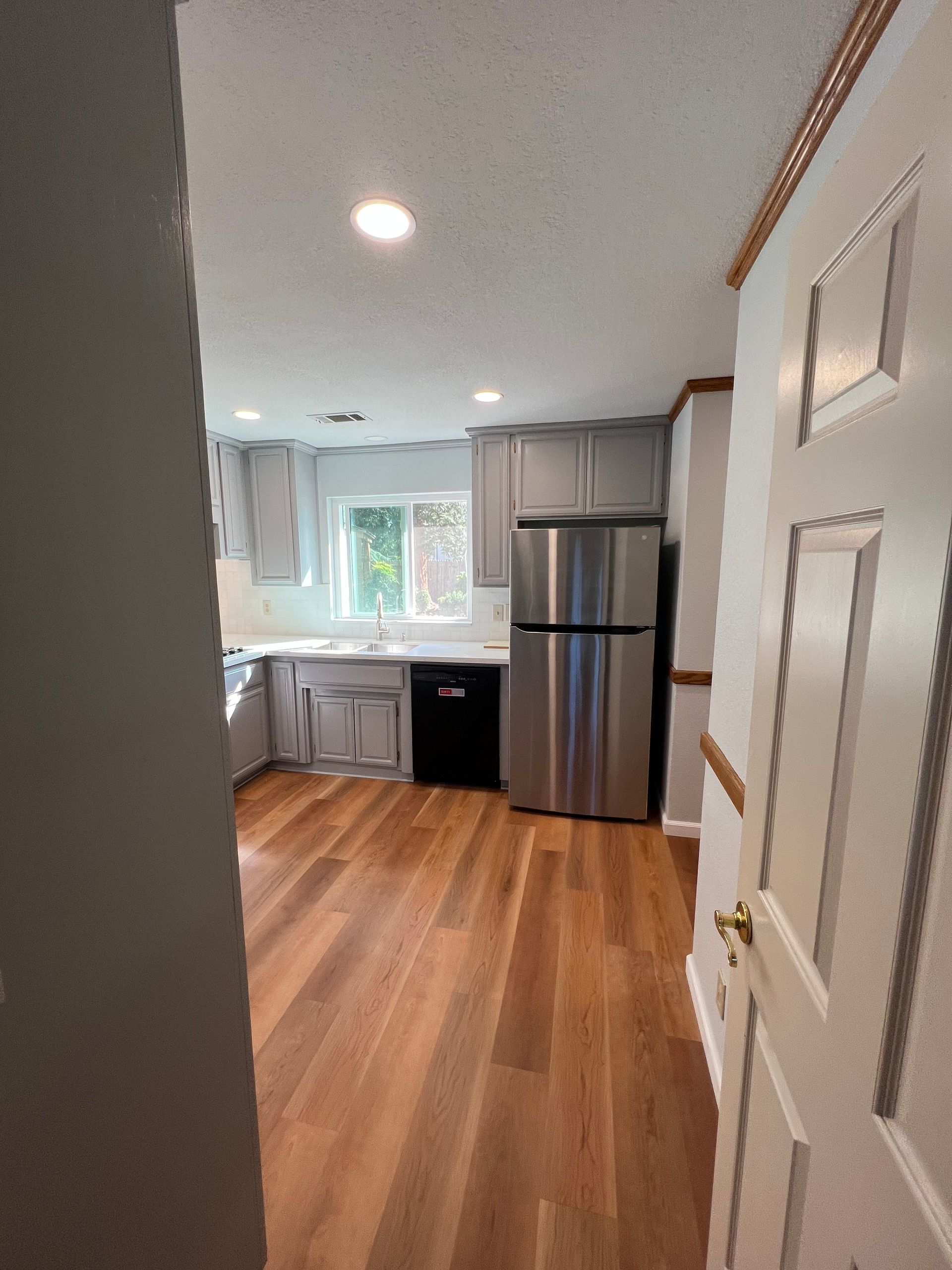Kitchen with gray cabinets, stainless steel refrigerator, and wooden floor. A door is visible on the right.