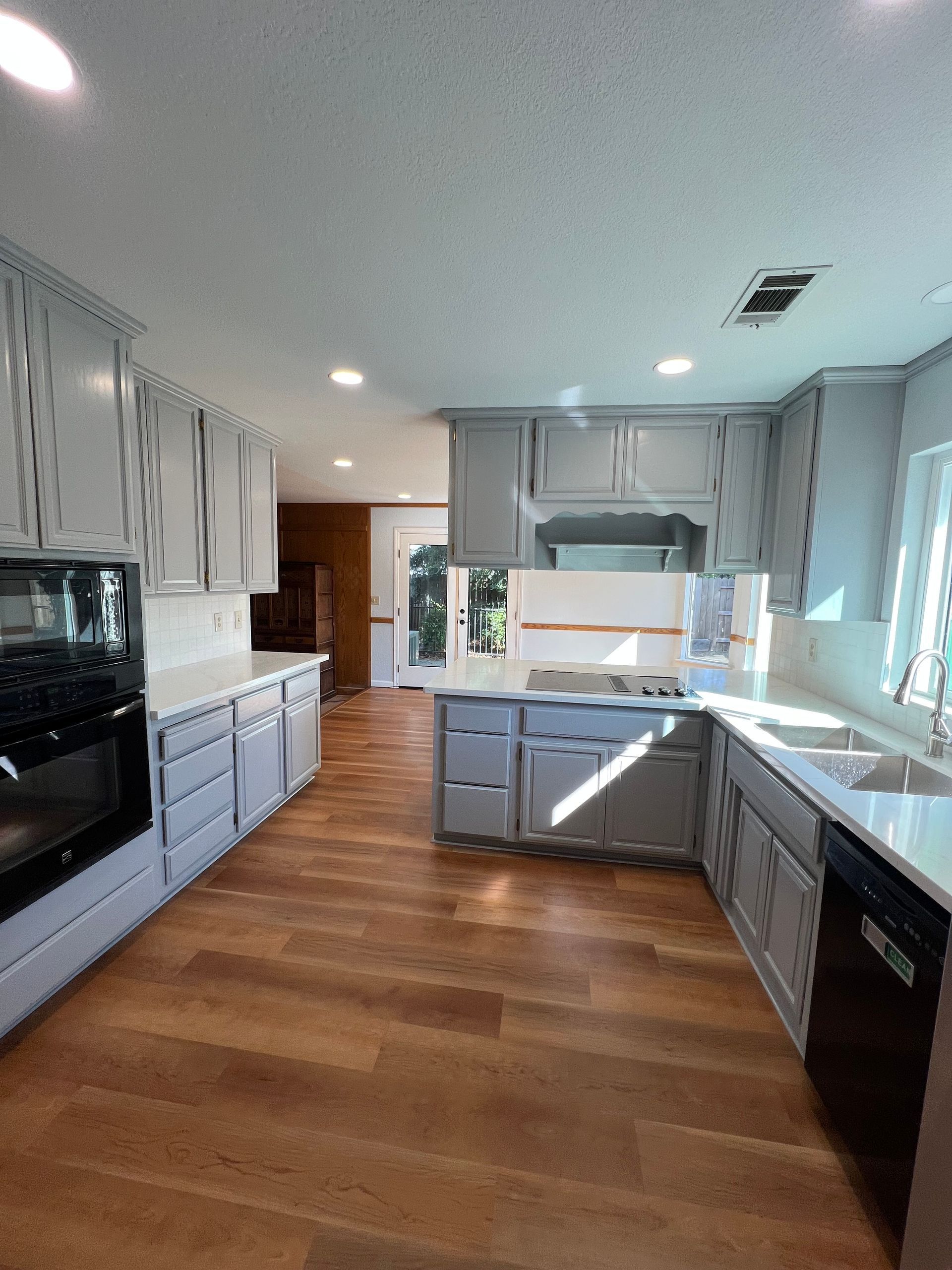 Spacious kitchen with gray cabinets, white countertops, and wooden floors. Sunlight streams in.