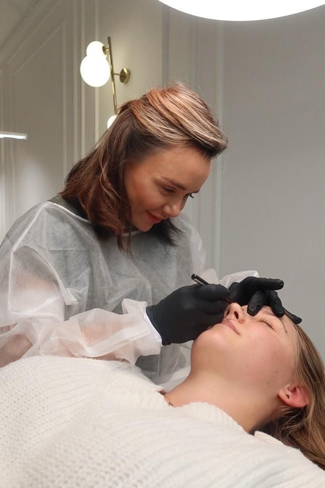 Woman in disposable gown drawing on another woman's eyebrow in a well-lit room.