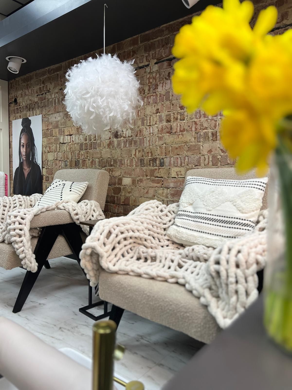 Two cozy armchairs with blankets, brick wall backdrop, and a fluffy white pendant light. Yellow flowers in the foreground.