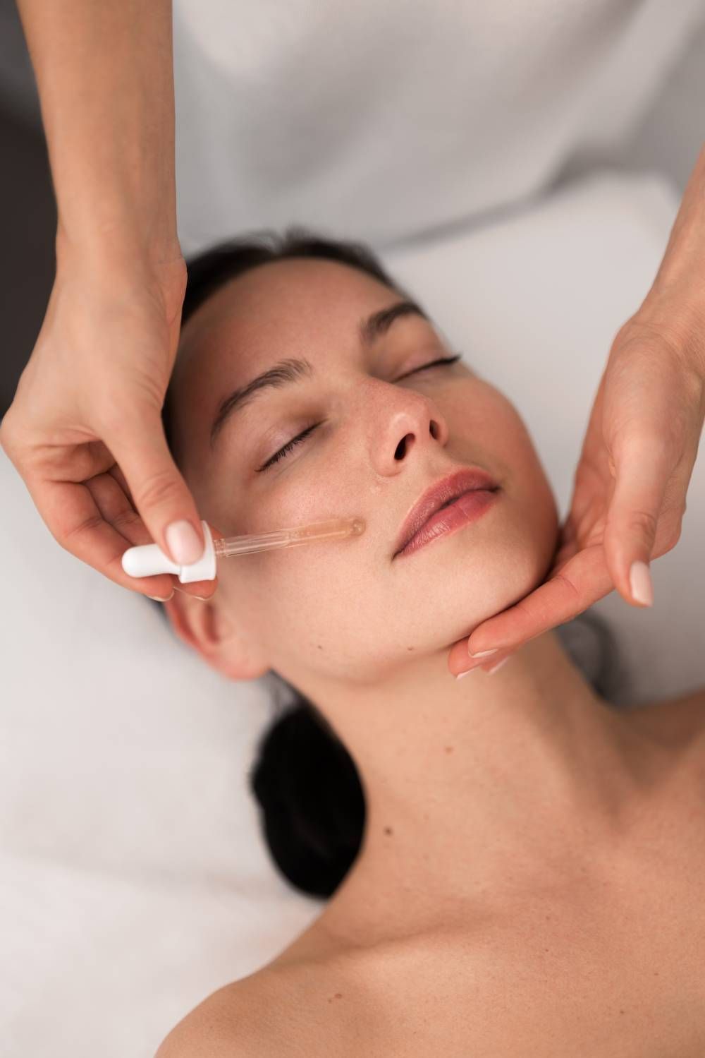 Person receiving facial treatment: hands applying serum to face with dropper, eyes closed.