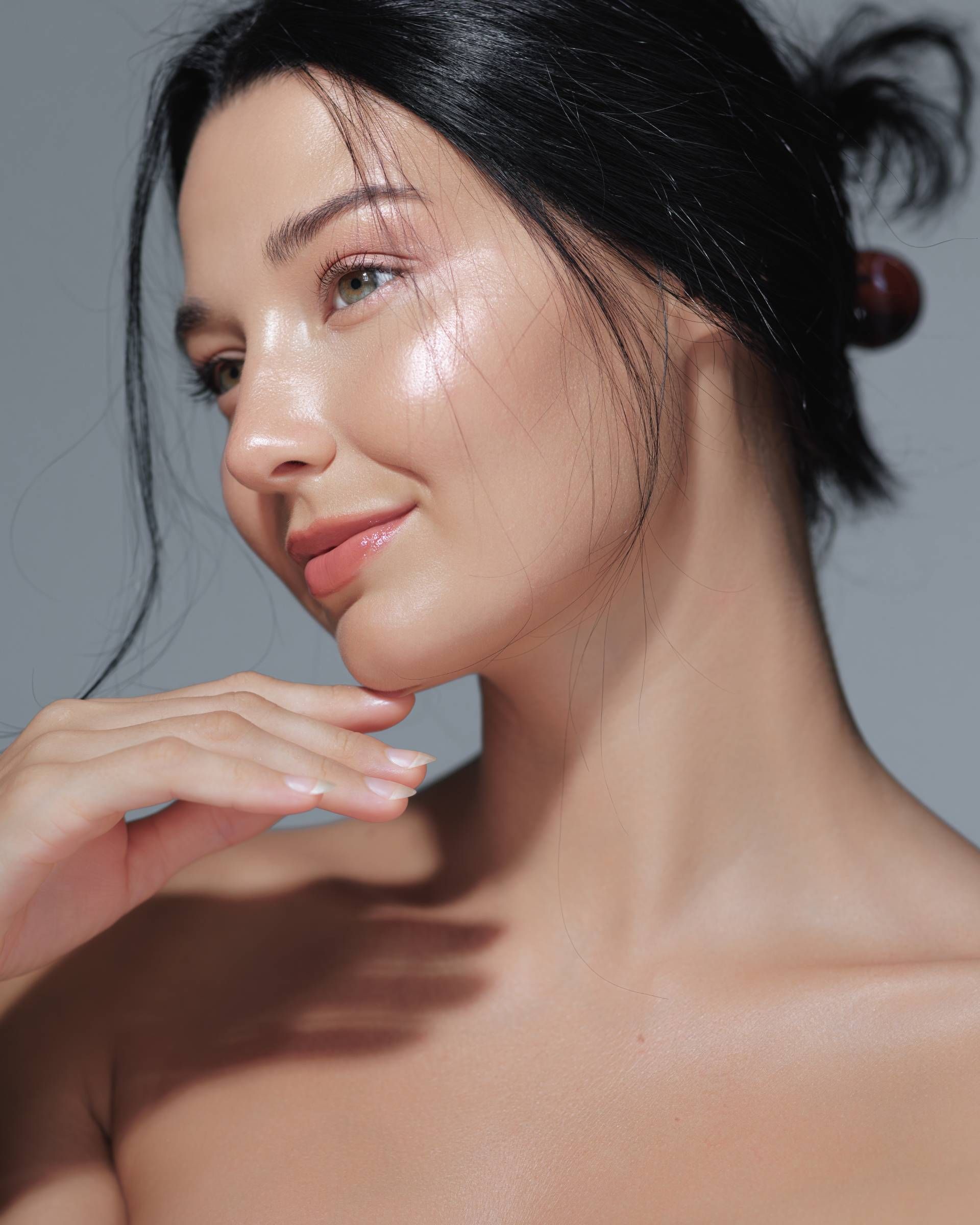 Woman with slicked-back dark hair, illuminated face, and hand under chin. Light makeup, soft smile.