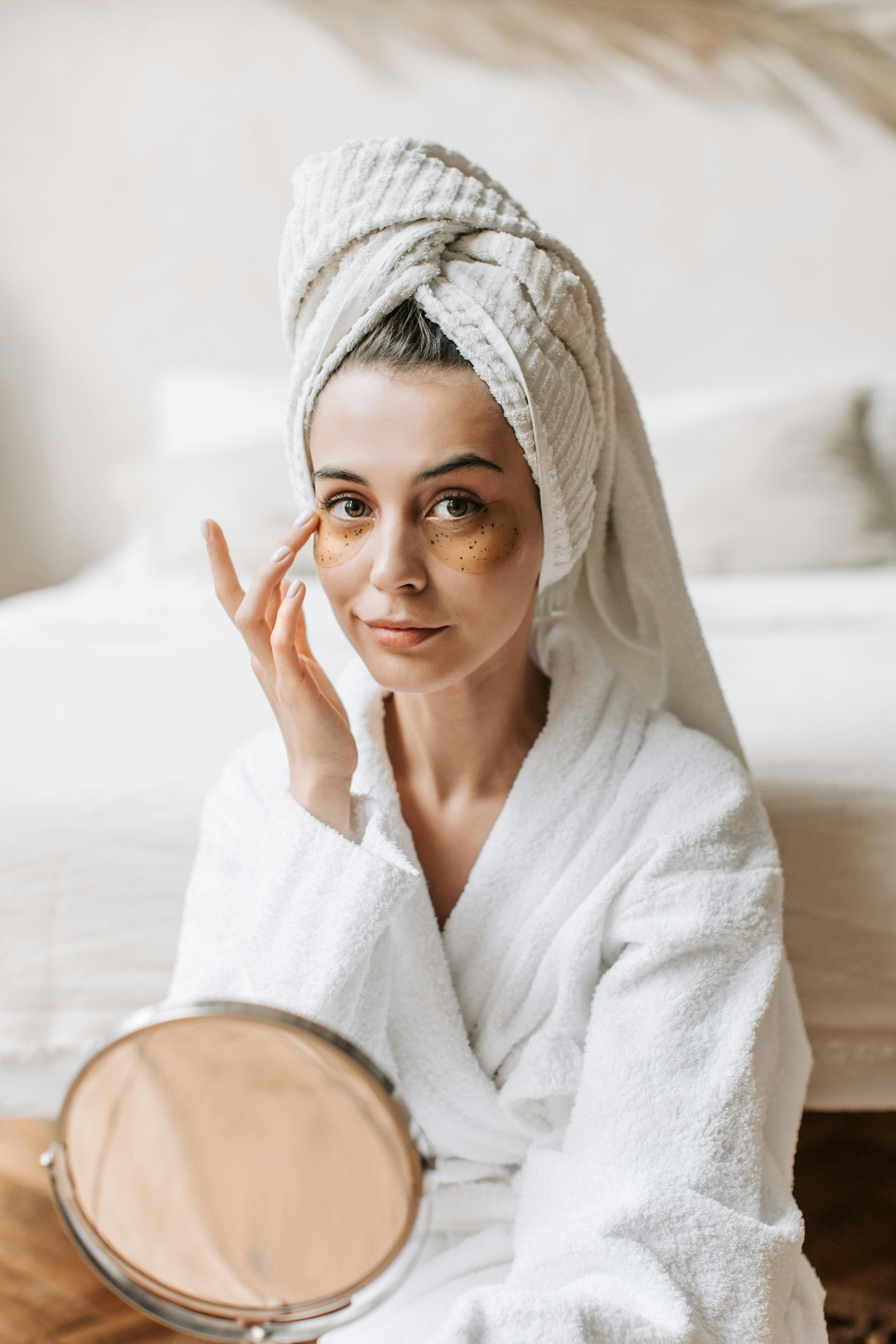 Woman in white robe and towel applies under-eye patches, looking at a mirror.