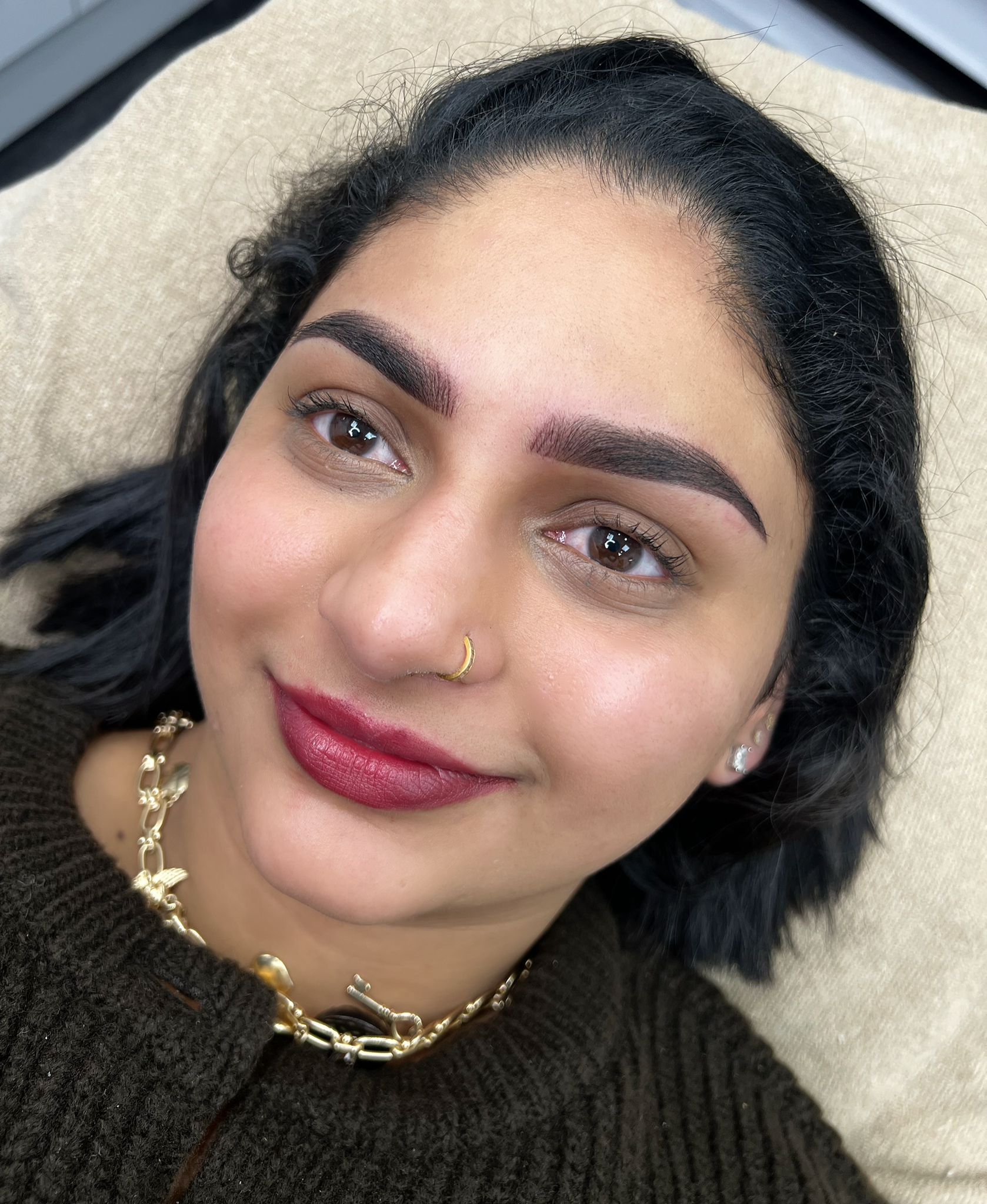 Woman with freshly done eyebrows, wearing a brown sweater, red lipstick, and a gold necklace, smiles at the camera.