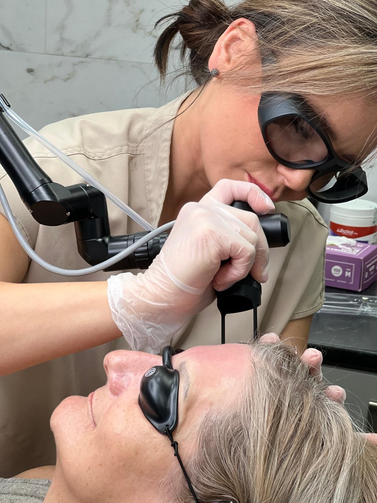 A person receiving laser skin treatment on the face. Practitioner holds device, both wear eye protection.