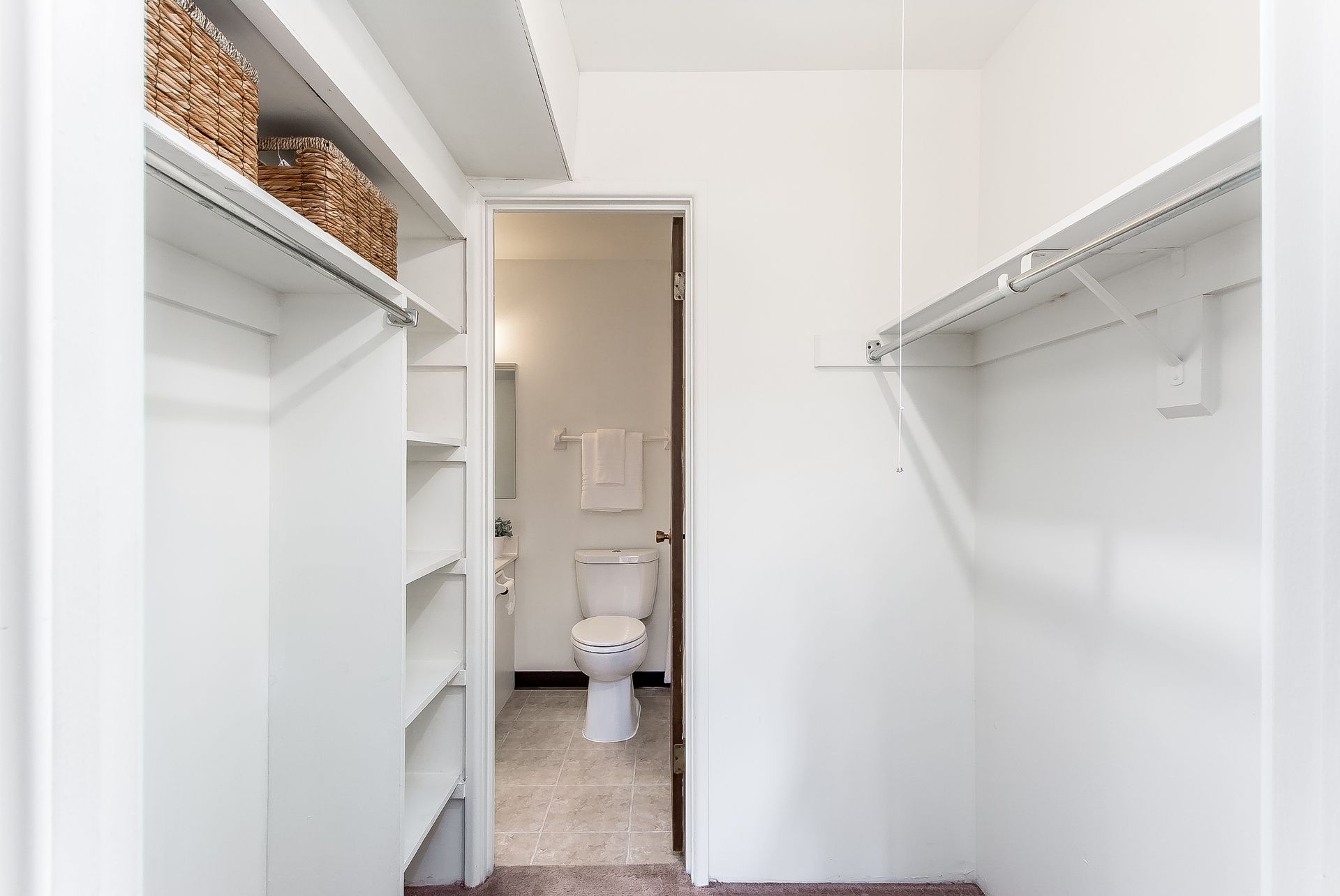 A walk in closet with a toilet and a bathroom in the background.