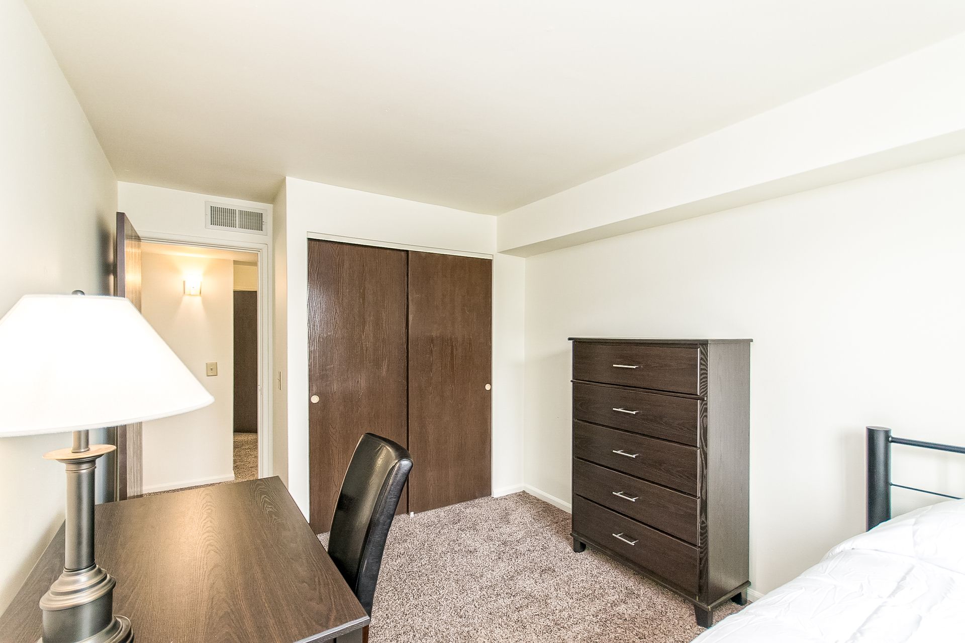 A bedroom with a bed , desk , lamp and dresser.