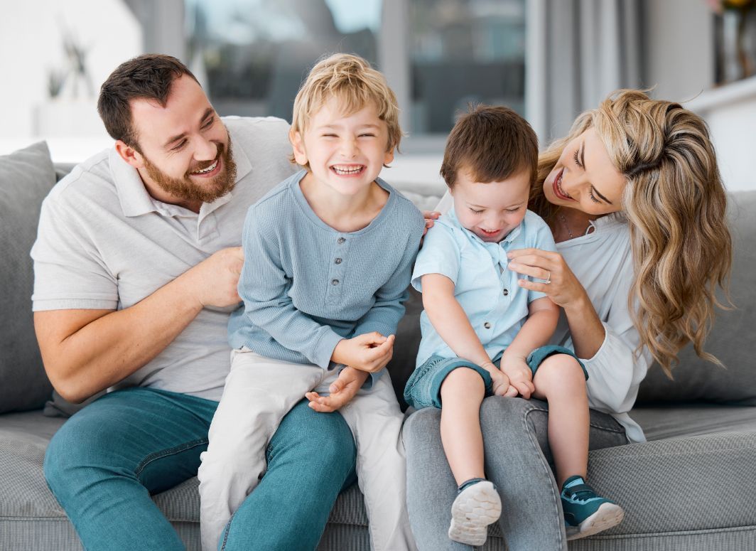 Family of four smiling on a gray couch; the parents with their two children.
