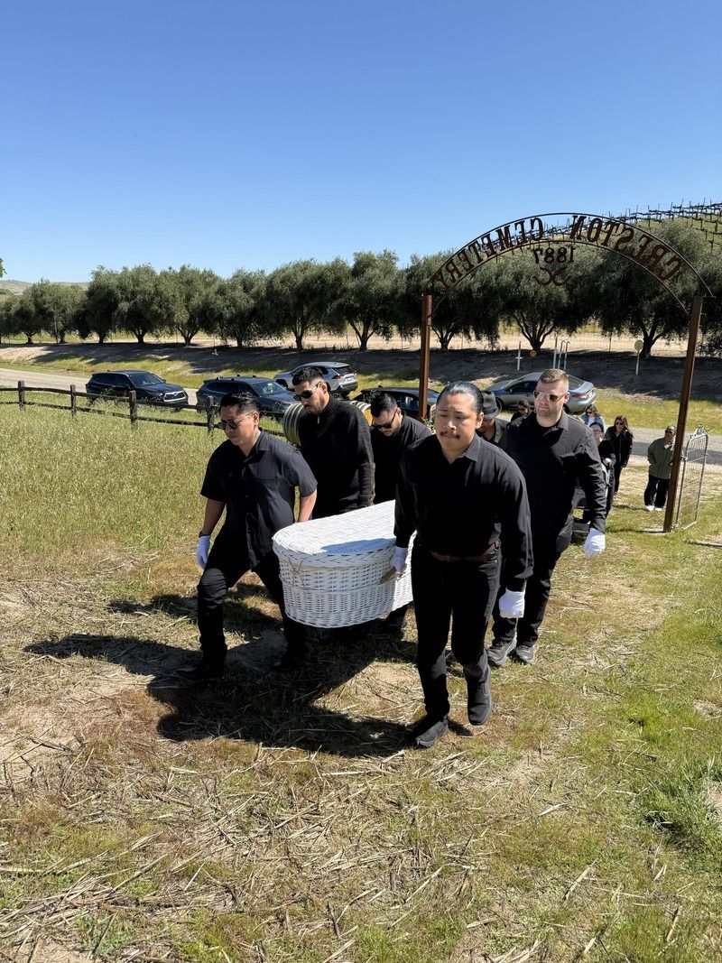 People carrying a white box through a grassy field with trees and parked cars in the background