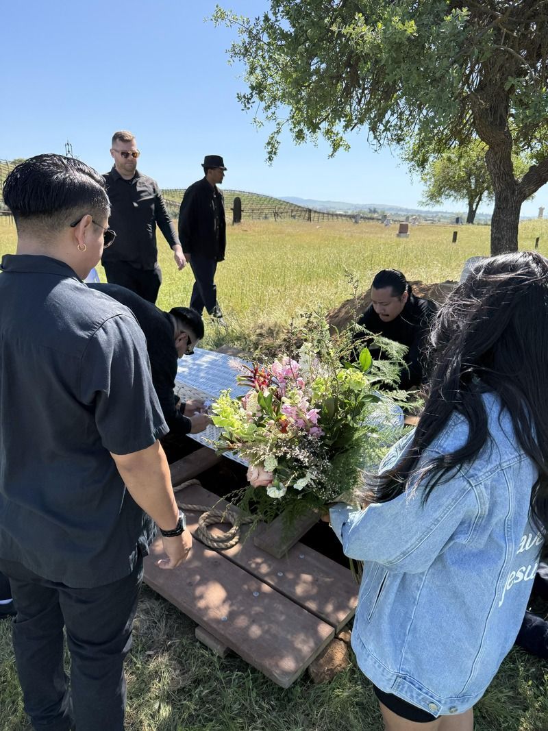 People gathered outdoors around a small memorial with flowers near a bench and tree in a grassy field.