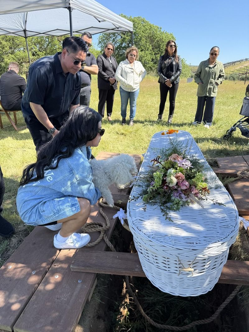 Mourners gather by a white casket with flowers at an outdoor graveside service.