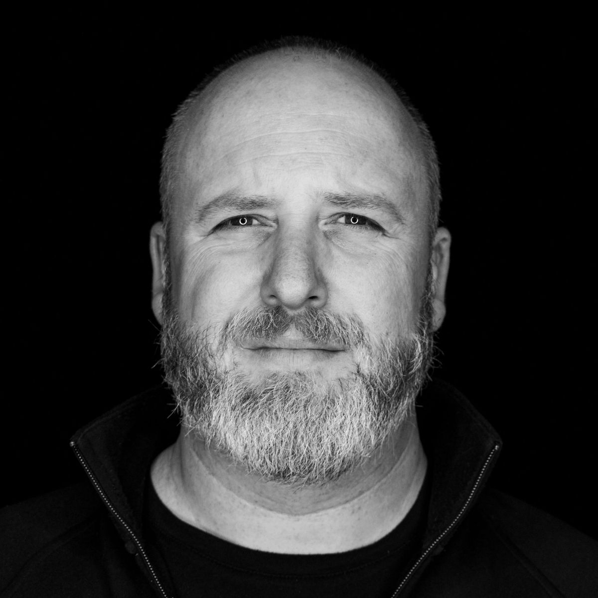 Man with a bald head and a gray beard, wearing a dark jacket, looking at the camera. Black background.