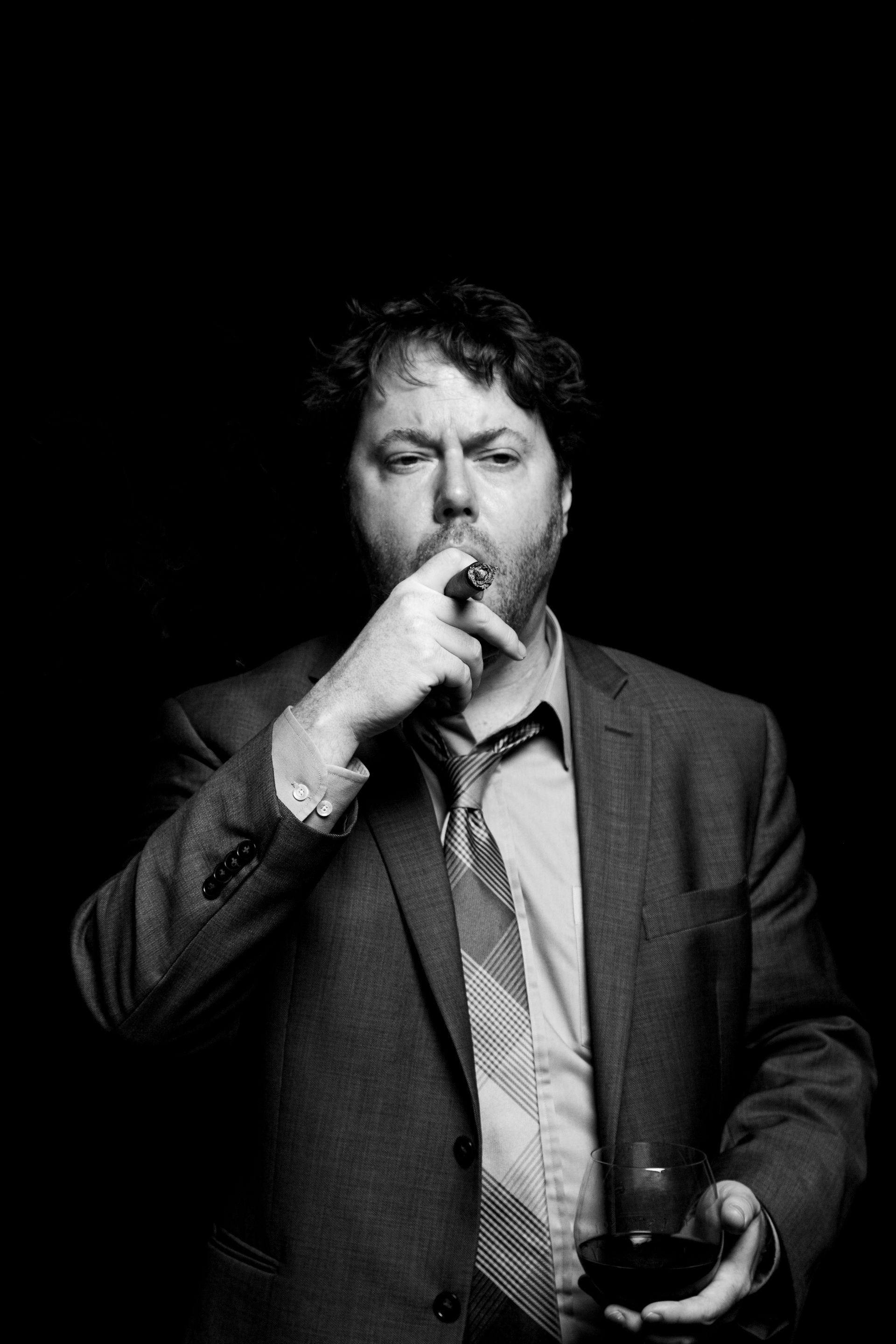 Man in suit smoking a cigar, holding a drink, dark background.
