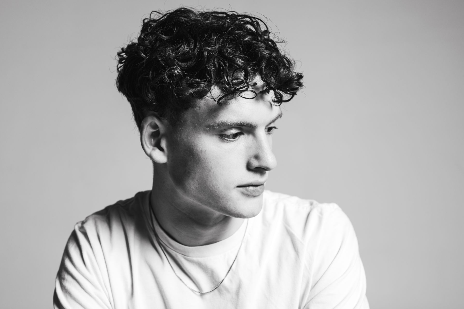 A young man with curly hair looks to the side in a studio, wearing a white t-shirt.