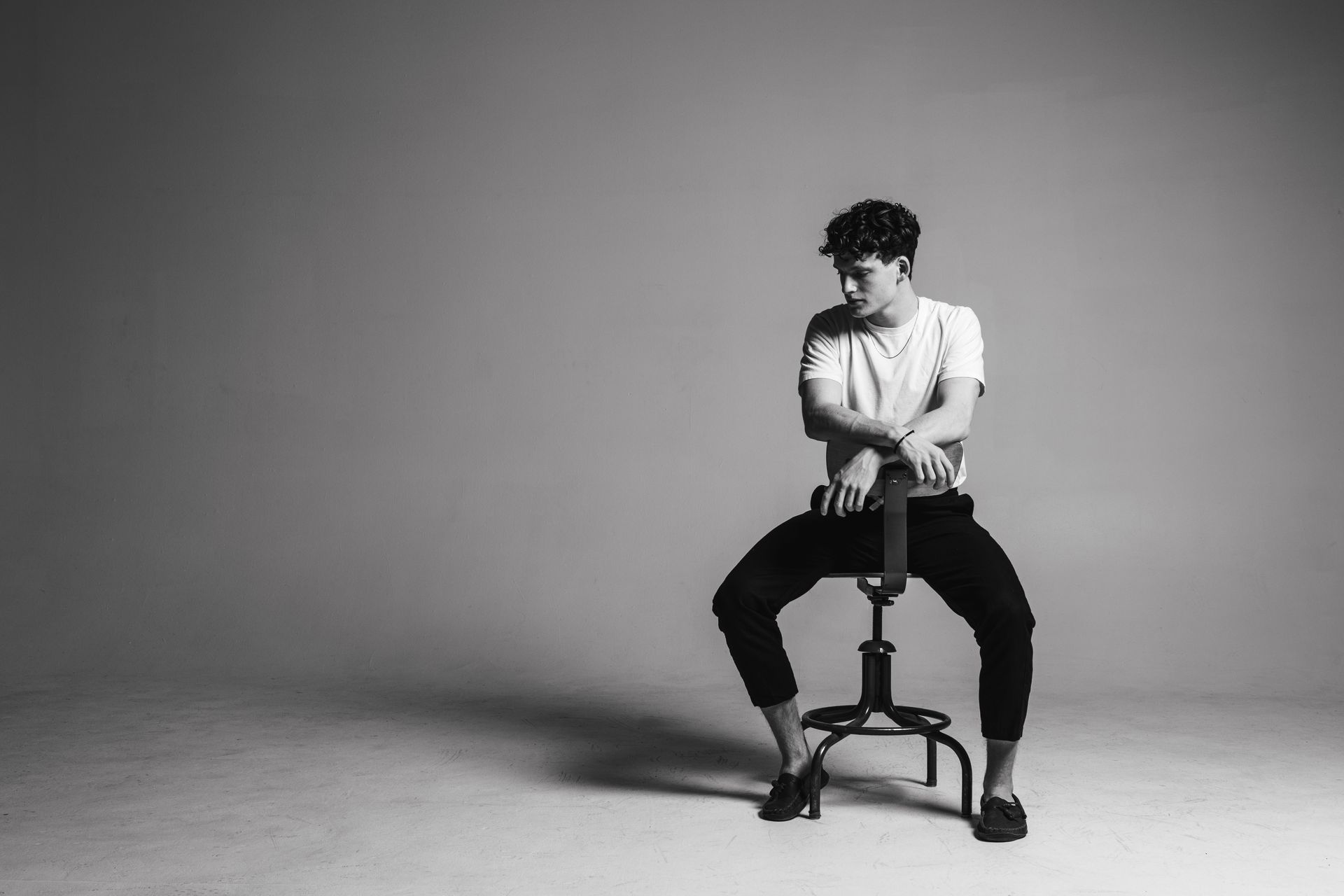 Man seated on stool, looking to the side with arms crossed over the stool back. Black and white photo.
