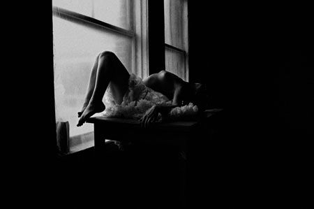 Woman in a white dress leans on a windowsill, silhouetted against a window. Black and white.