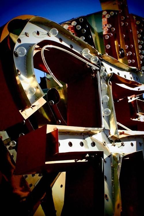 Close-up of a corroded neon sign frame in the sun. Metallic, gold, and red hues with a bright blue sky.