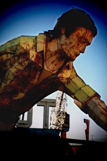 Giant statue of a man reaching down; green plaid shirt, weathered appearance, set against a daytime sky, with neon sign.