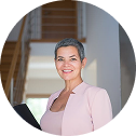 Woman with short gray hair wearing a pink blazer, smiling in a home interior.