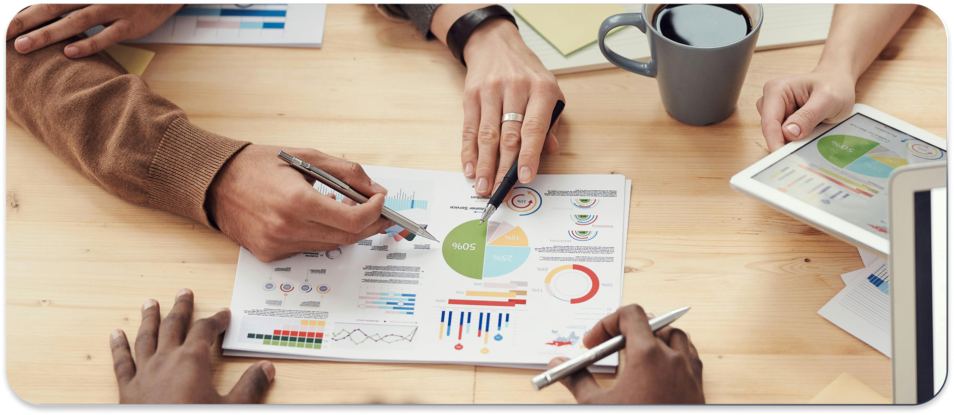 People are gathered around a table, analyzing charts and graphs with pens. There is a coffee mug and a tablet on the table.