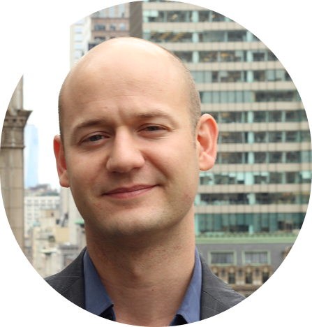 Man with shaved head in front of city buildings, smiling and looking at the camera.
