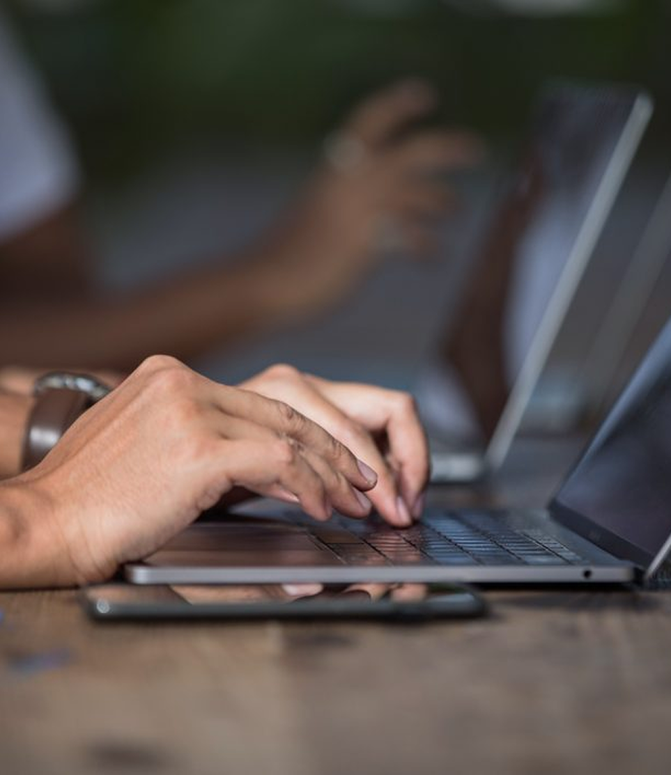 Hands typing on a laptop at a wooden table, with a blurred person and phone in the background