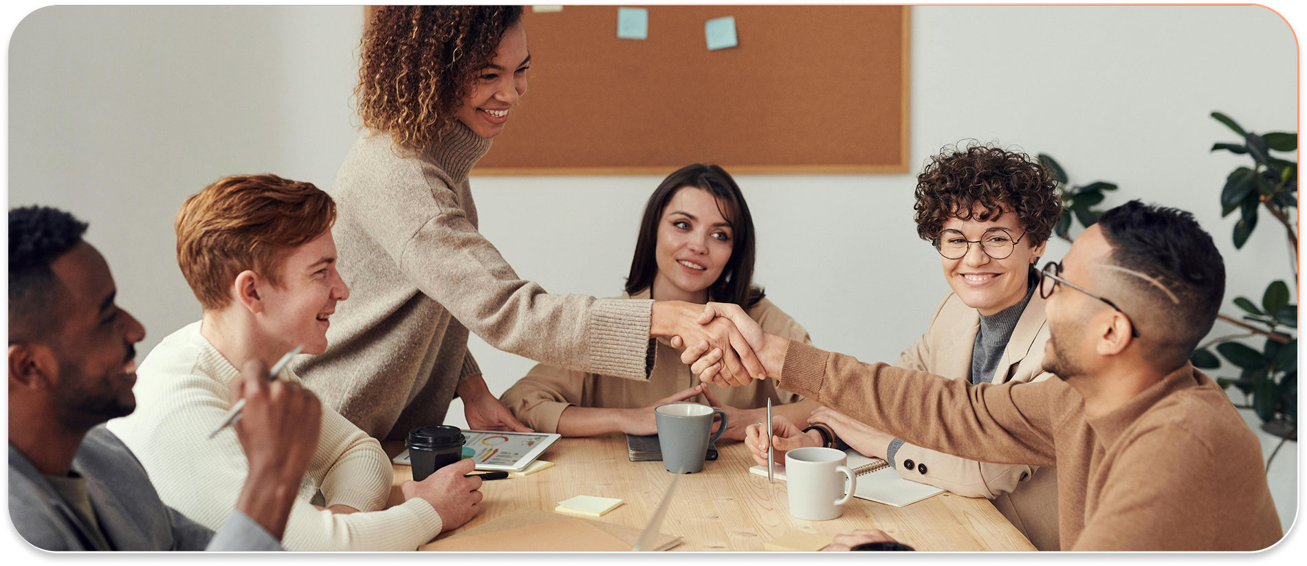 People in a business meeting shaking hands to greet each other. They are sitting at a table in a bright room with a bulletin board.
