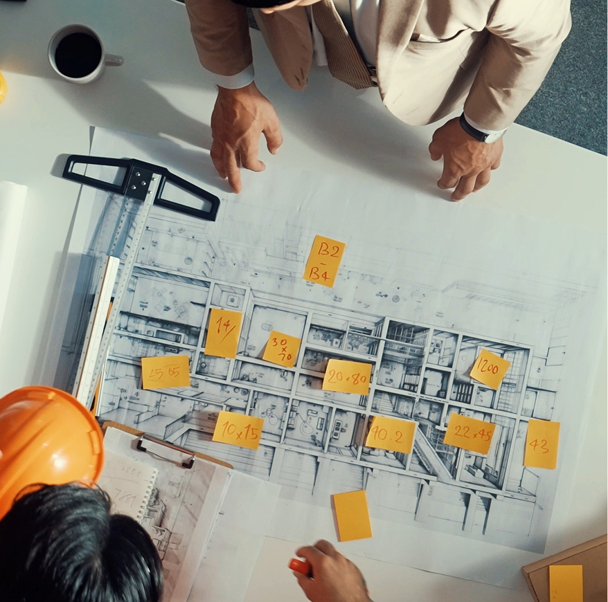 Two men shaking hands at a construction site; one in a suit and white hard hat, the other in work attire and yellow hard hat.