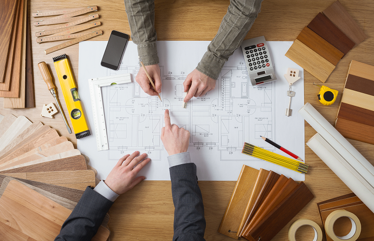 Two people reviewing blueprints, surrounded by wood samples, tools, and a calculator on a desk.