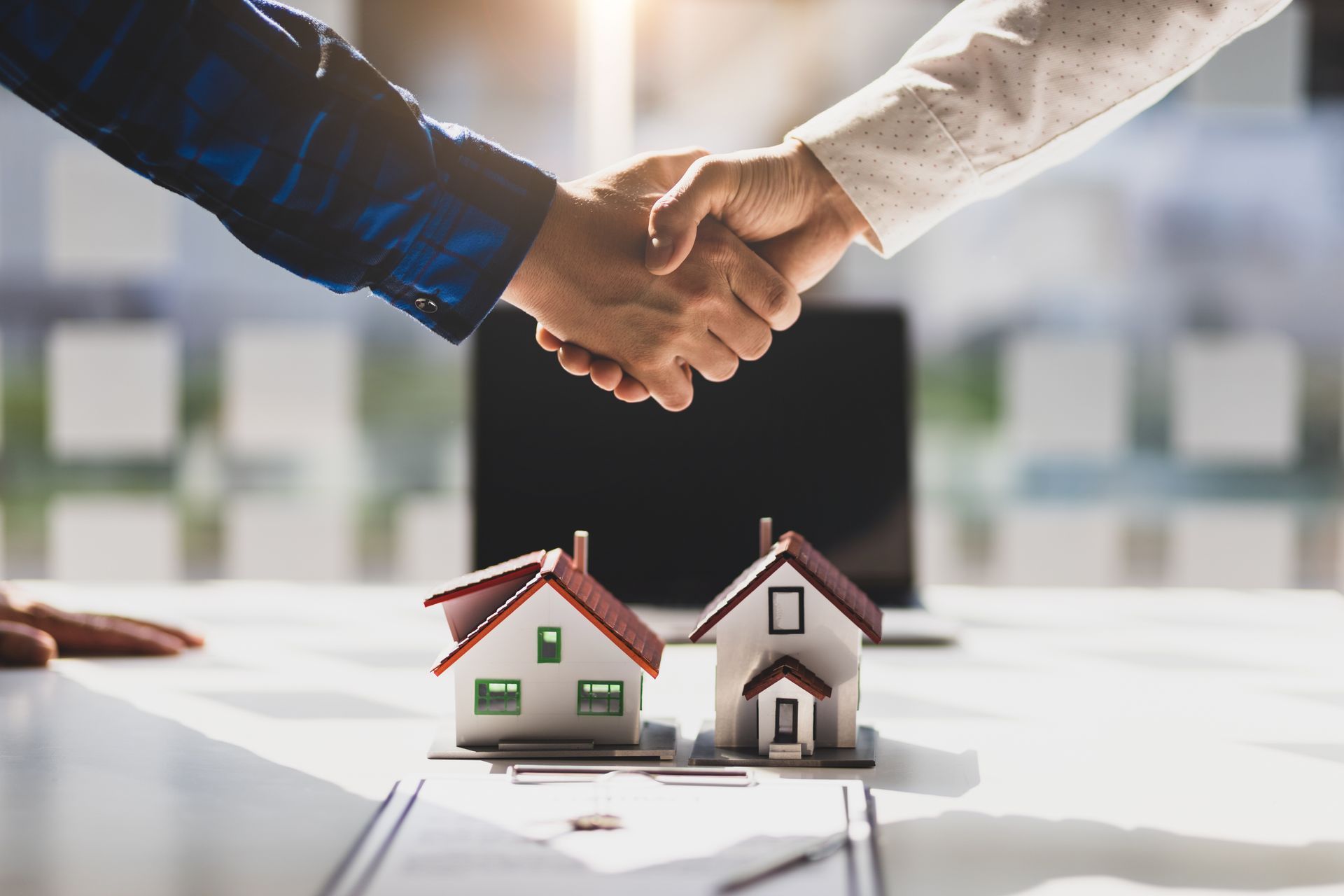 Two people shaking hands over miniature houses and paperwork, real estate deal.