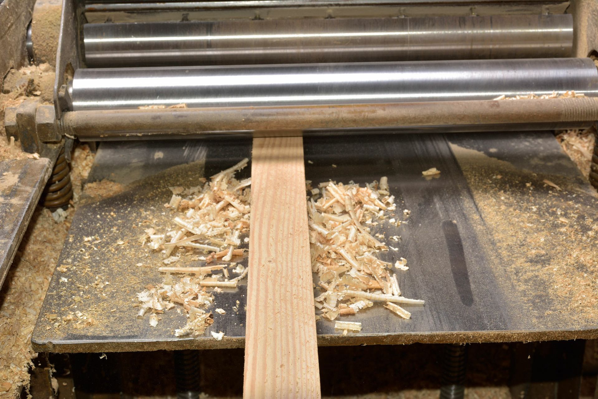 A wood plank being planed by a machine, producing wood shavings.