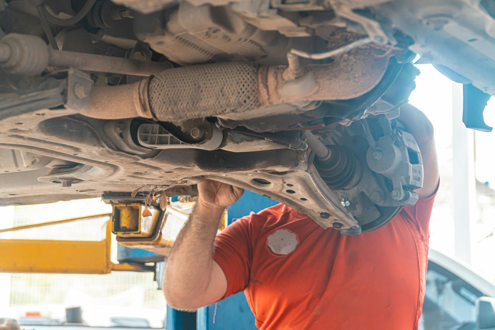 Mechanic in red shirt working under a car on a lift. Exhaust pipe and suspension visible.