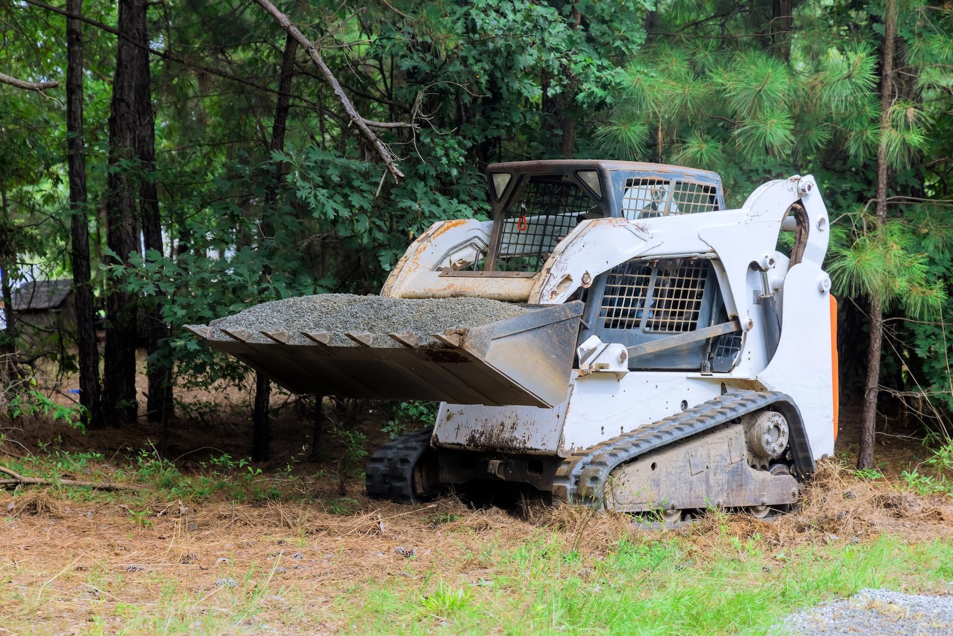White Bobcat with a toothed bucket, scooping gravel in a wooded area.
