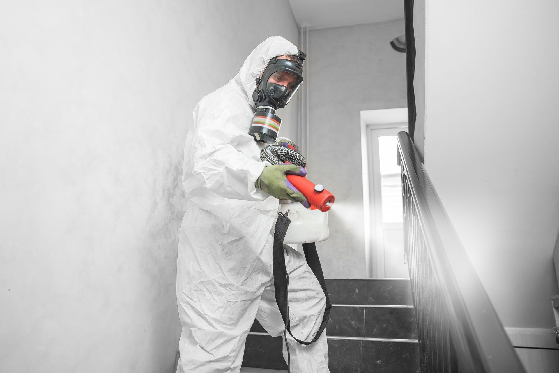 A professional in a white hazmat suit and respirator sprays disinfectant on a staircase.