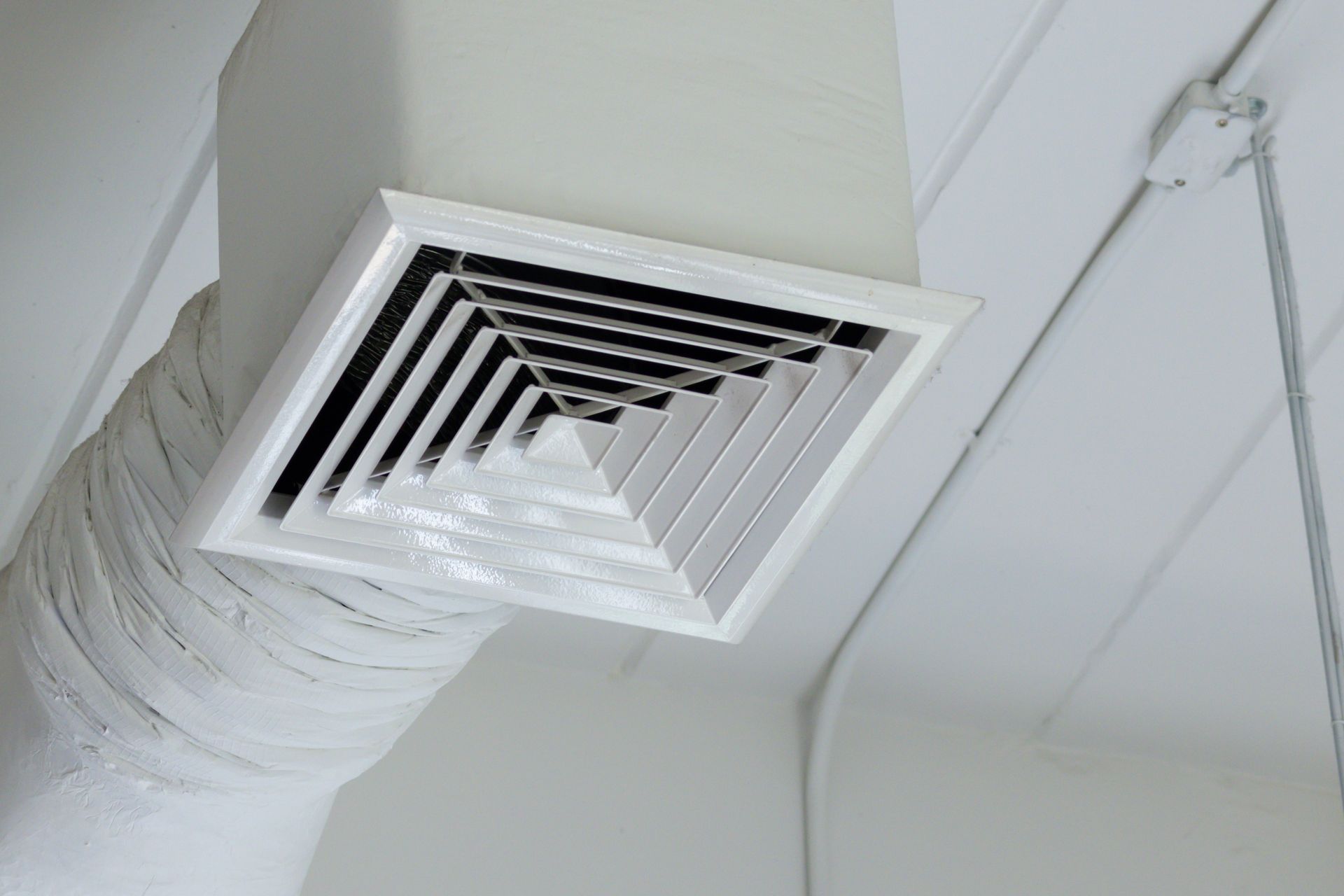 A square white ceiling air vent with an attached flexible duct, set against a plain white ceiling.