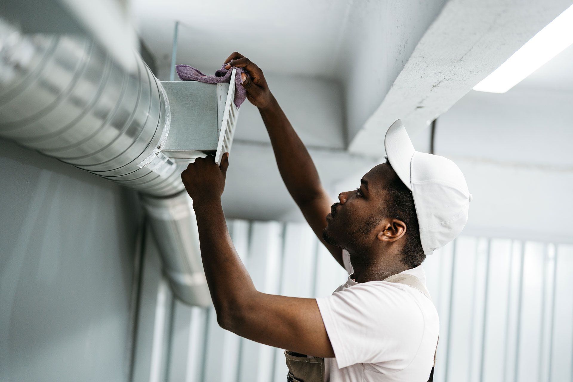 A technician in a white uniform and cap cleans or services a metallic ceiling air vent.