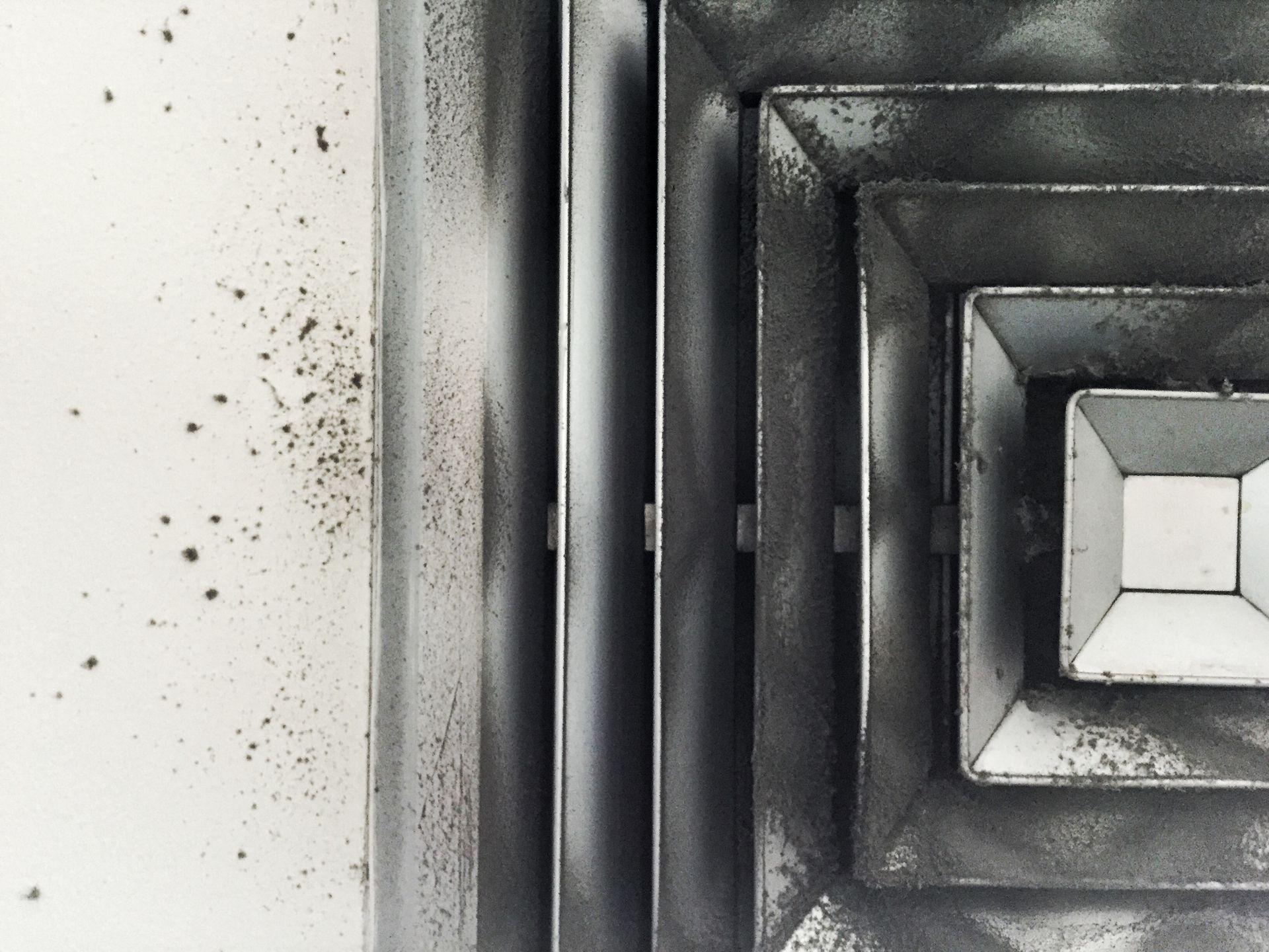 A close-up, high-angle view of a dirty, square metal air vent on a ceiling with dark dust streaks on the surrounding wall.