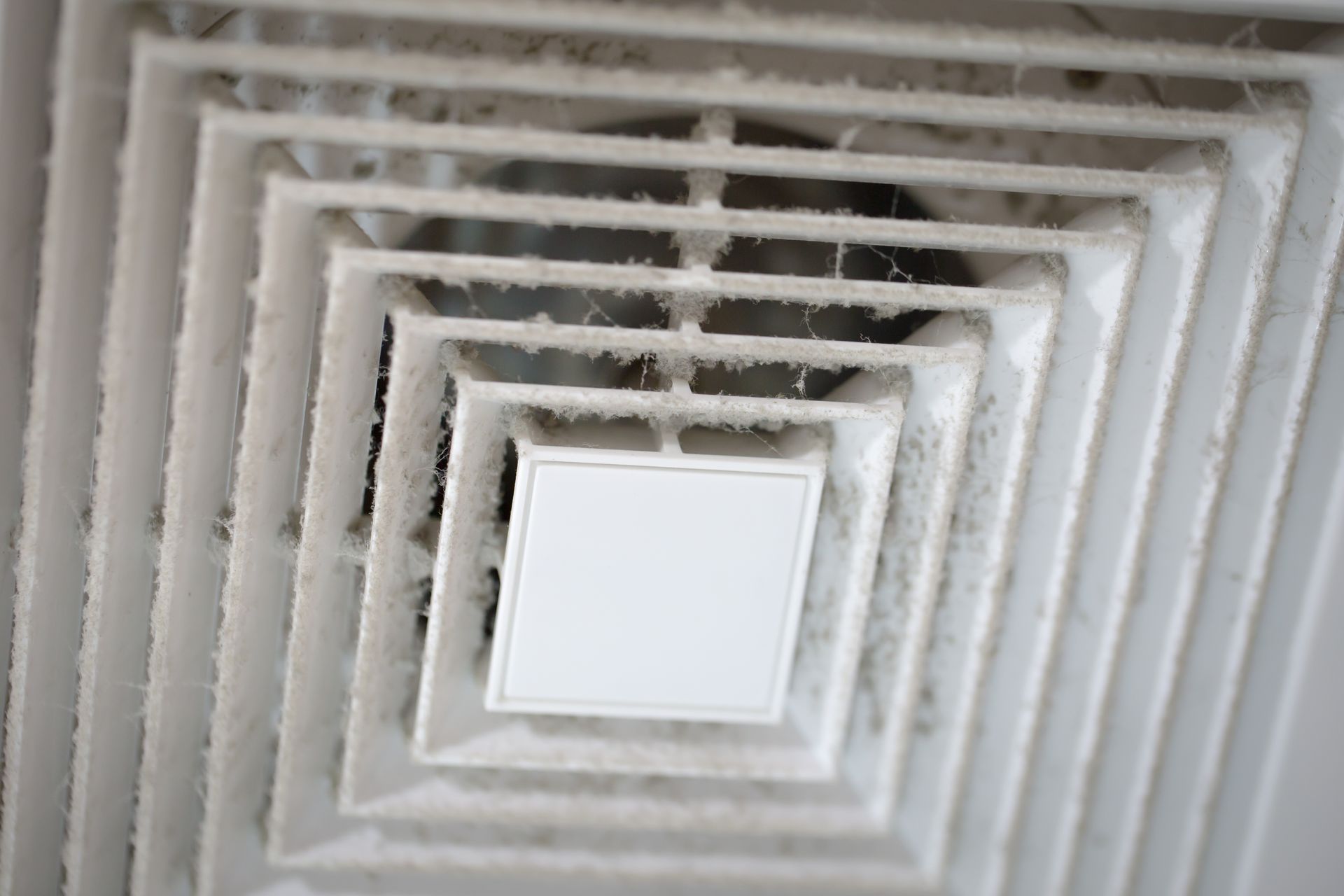 A close-up, angled view of a white, square ceiling air vent covered in a thick layer of grey dust and lint.
